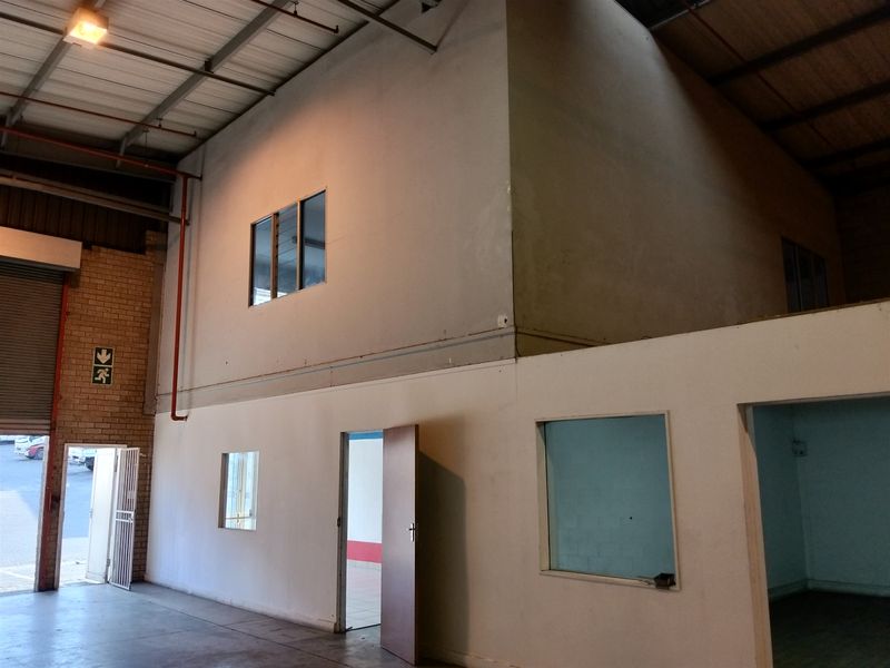 Industrial warehouse space featuring a mezzanine level, exposed steel roof trusses, fire sprinkler system, and internal office partitioning with roller shutter access.
