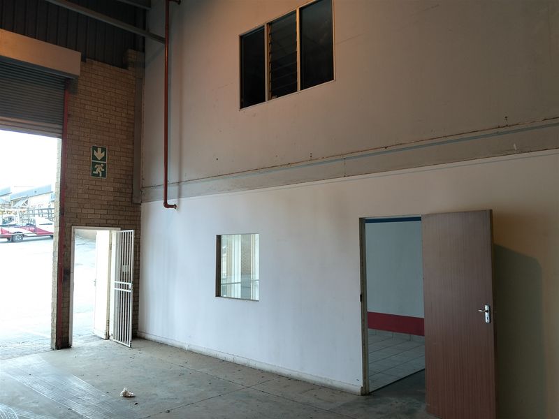 Industrial warehouse space featuring a large roller shutter door, high ceilings with fire suppression piping, and an integrated office or utility room.