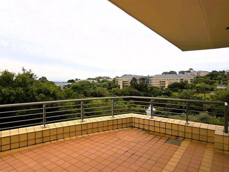 A spacious outdoor balcony featuring tiled flooring and metal railings, offering expansive views of the surrounding office park and greenery.