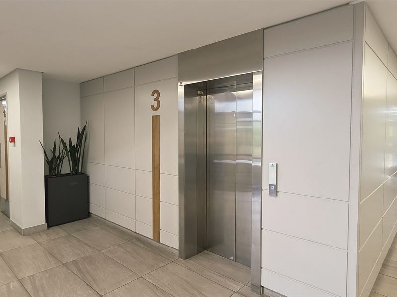 Modern office building hallway featuring a stainless steel elevator, tiled flooring, and recessed linear lighting with glass-paneled access doors.