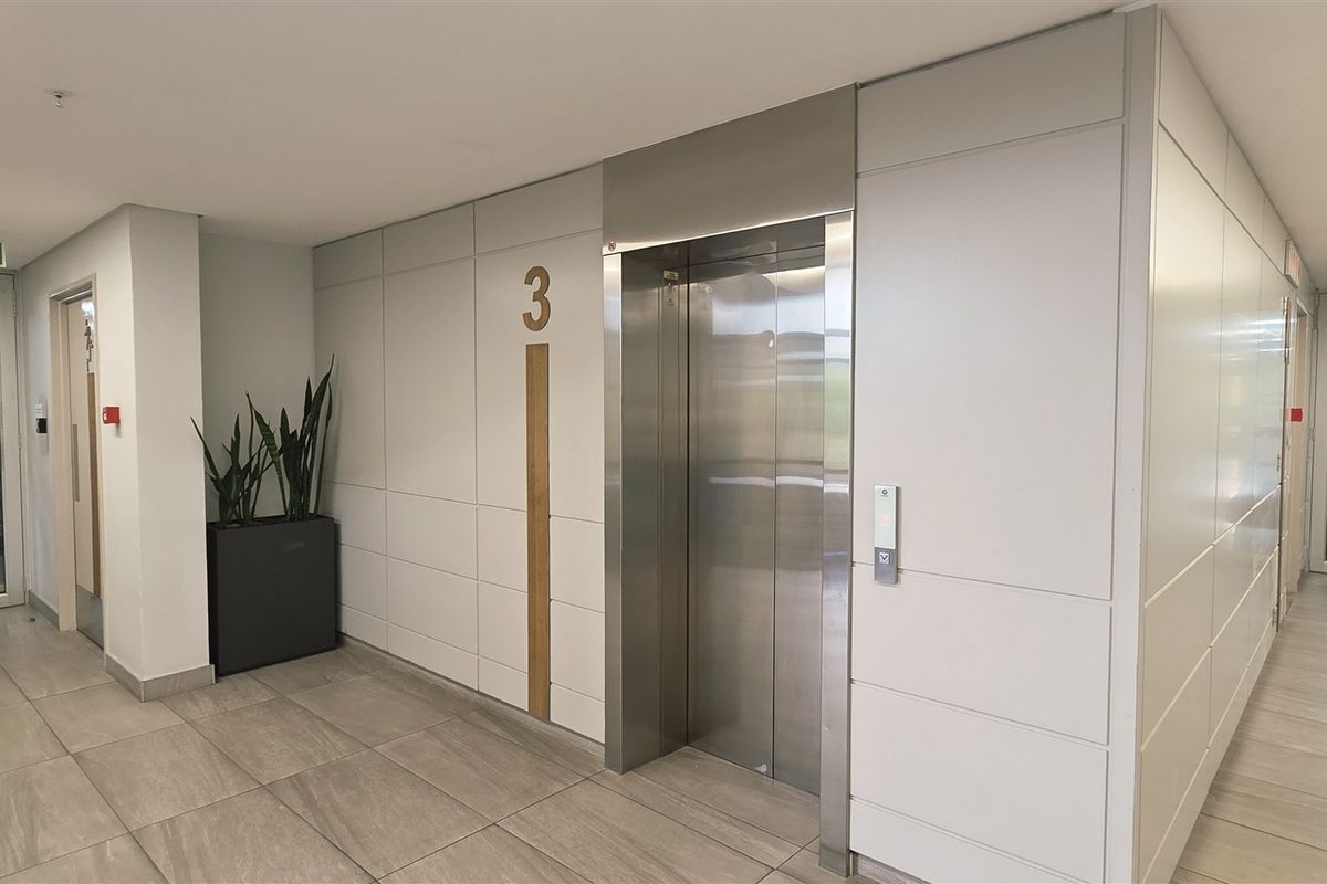 Modern office building hallway featuring a stainless steel elevator, tiled flooring, and recessed linear lighting with glass-paneled access doors.