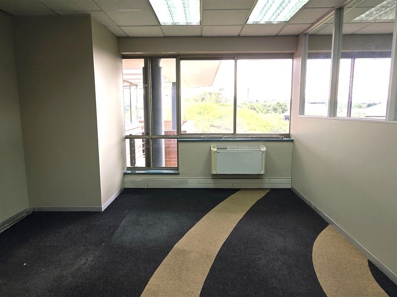 An empty office space featuring large windows, a wall-mounted air conditioning unit, suspended ceiling, and patterned carpeting with glass partition walls.