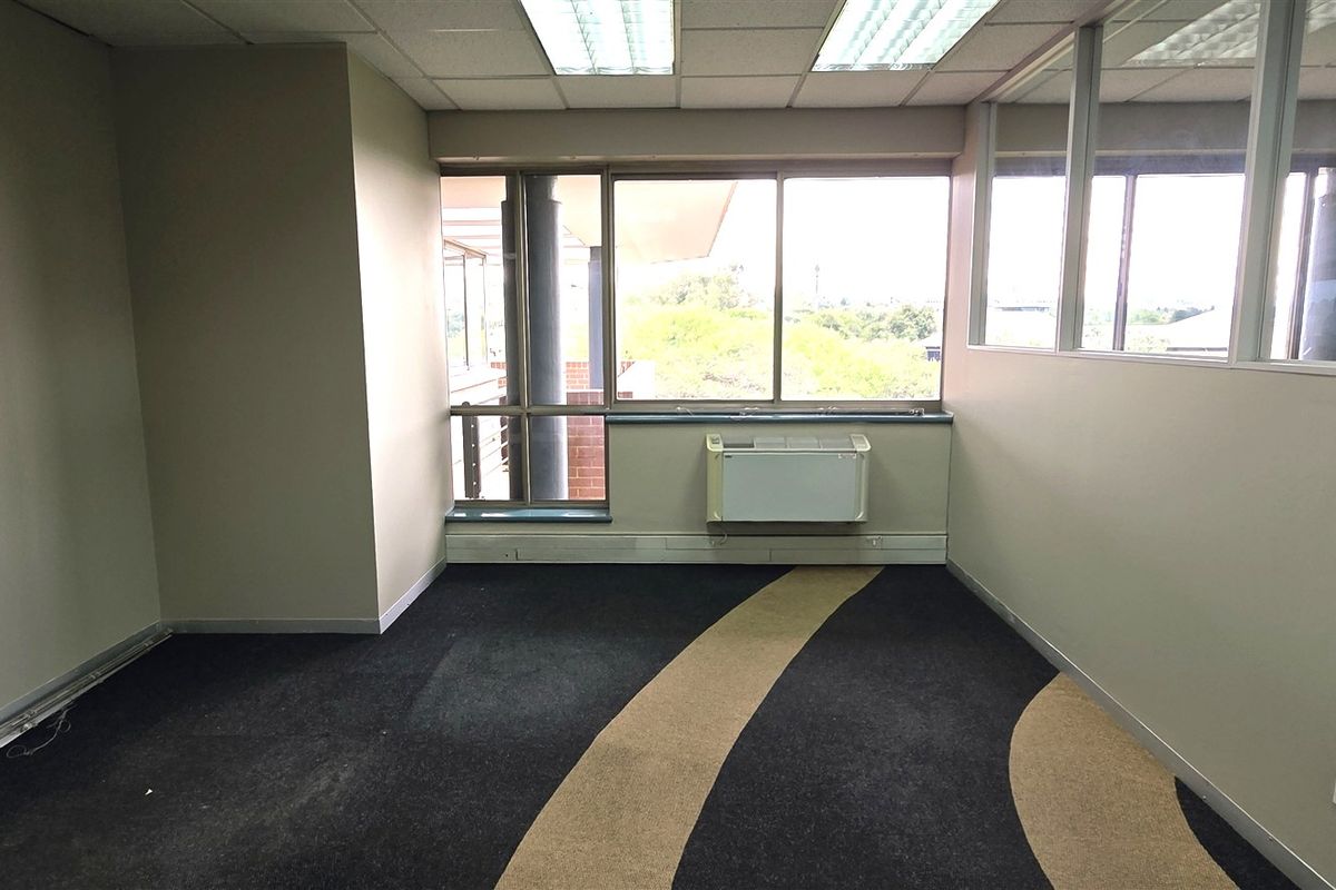 An empty office space featuring large windows, a wall-mounted air conditioning unit, suspended ceiling, and patterned carpeting with glass partition walls.