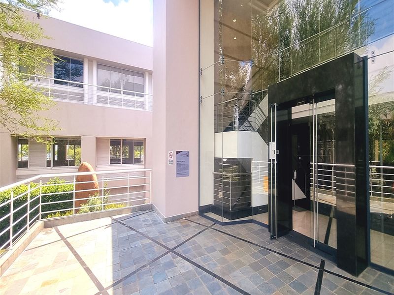 Modern office building entrance featuring a glass facade, stone tiled patio, and a prominent glass-enclosed entryway with a visible internal staircase.