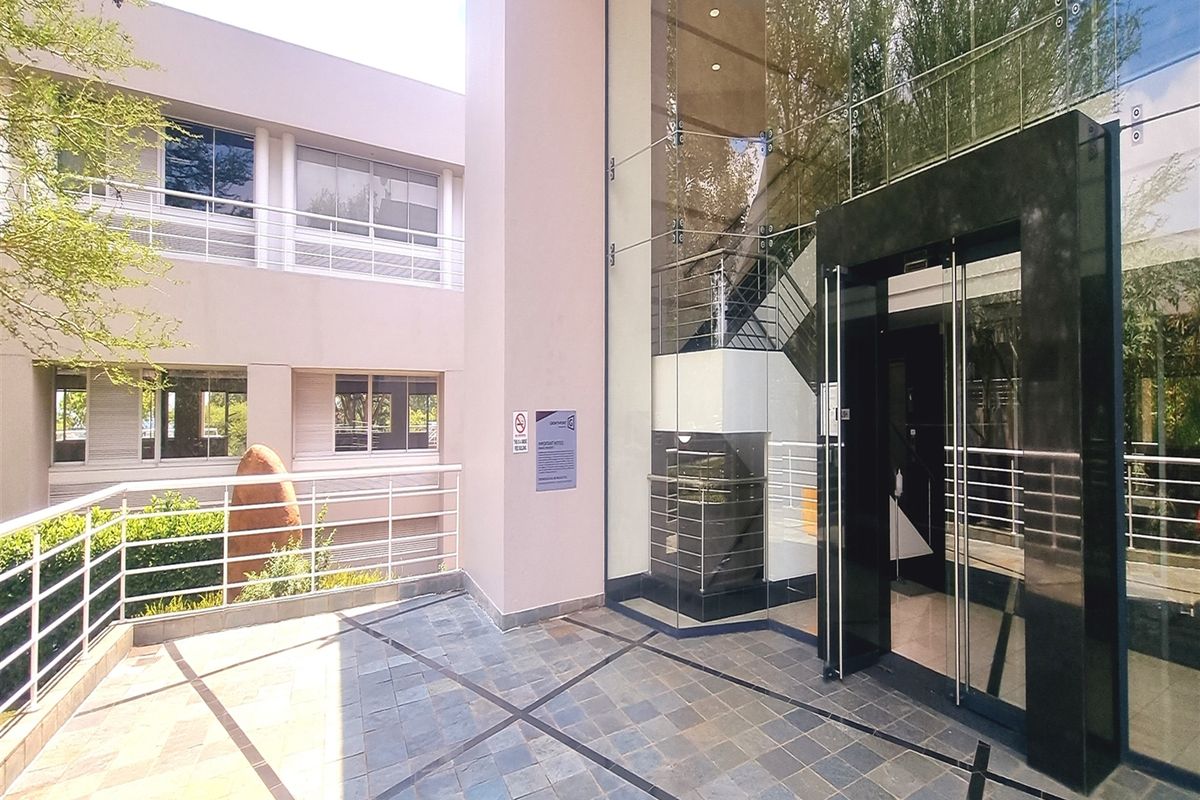 Modern office building entrance featuring a glass facade, stone tiled patio, and a prominent glass-enclosed entryway with a visible internal staircase.