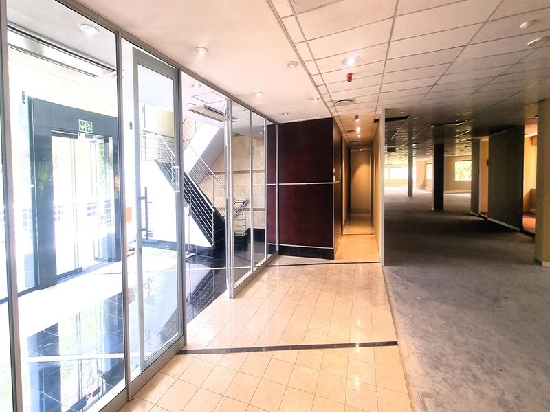Spacious office hallway featuring glass partitions, tiled flooring, and a suspended ceiling, leading to an open-plan office area and staircase.