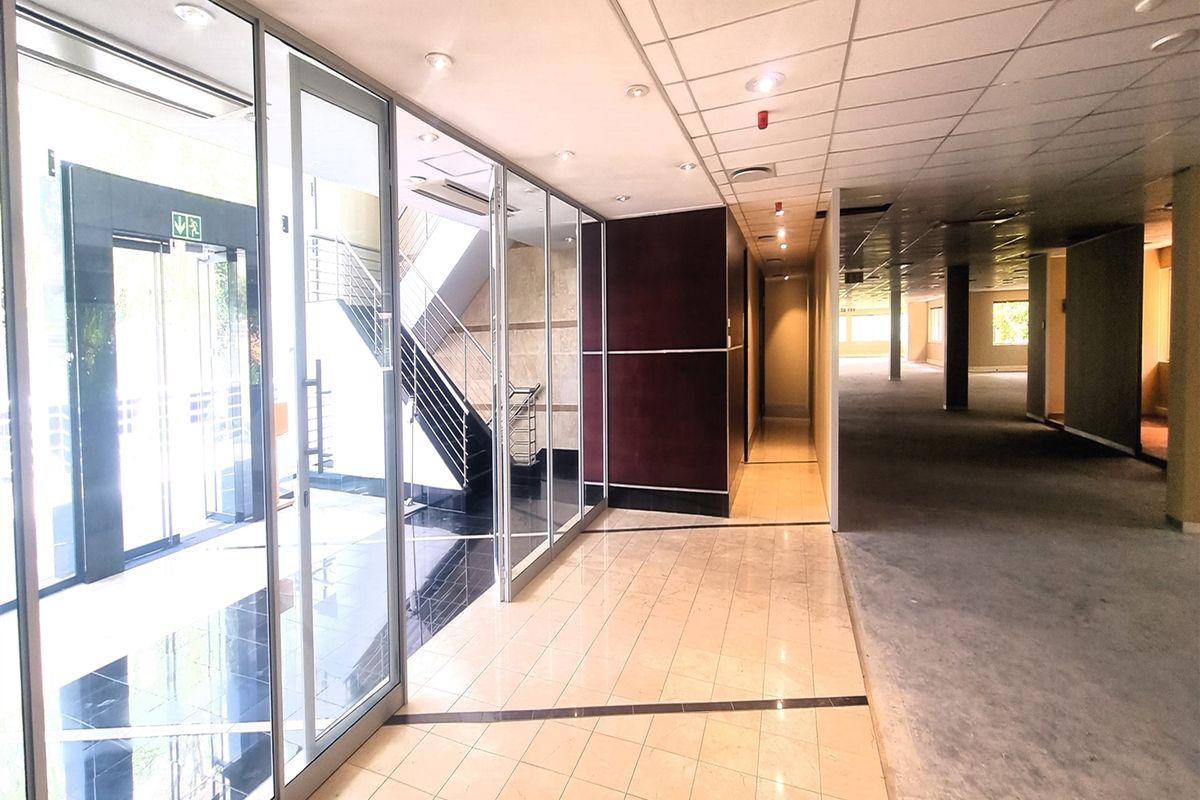 Spacious office hallway featuring glass partitions, tiled flooring, and a suspended ceiling, leading to an open-plan office area and staircase.