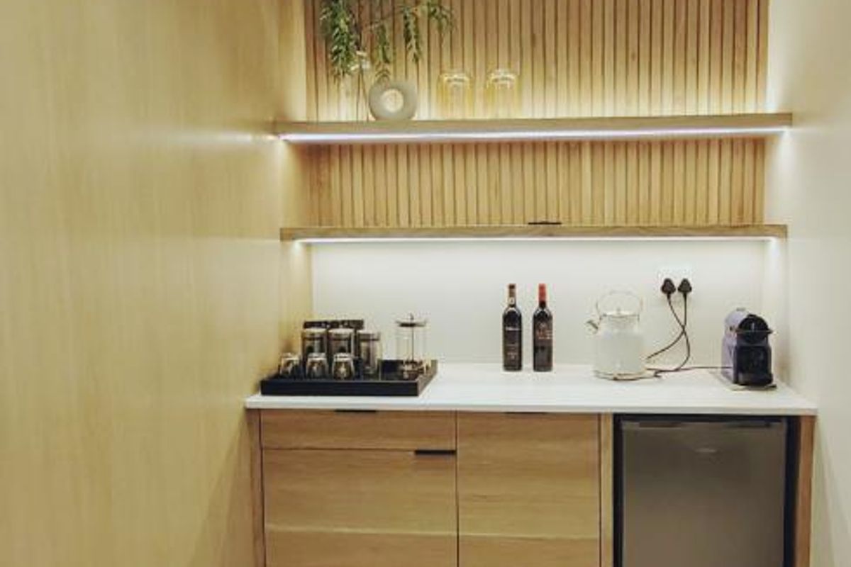 A modern kitchenette area featuring light wood cabinetry, integrated under-counter fridge space, stone countertop, and decorative slatted timber wall paneling with integrated LED lighting.