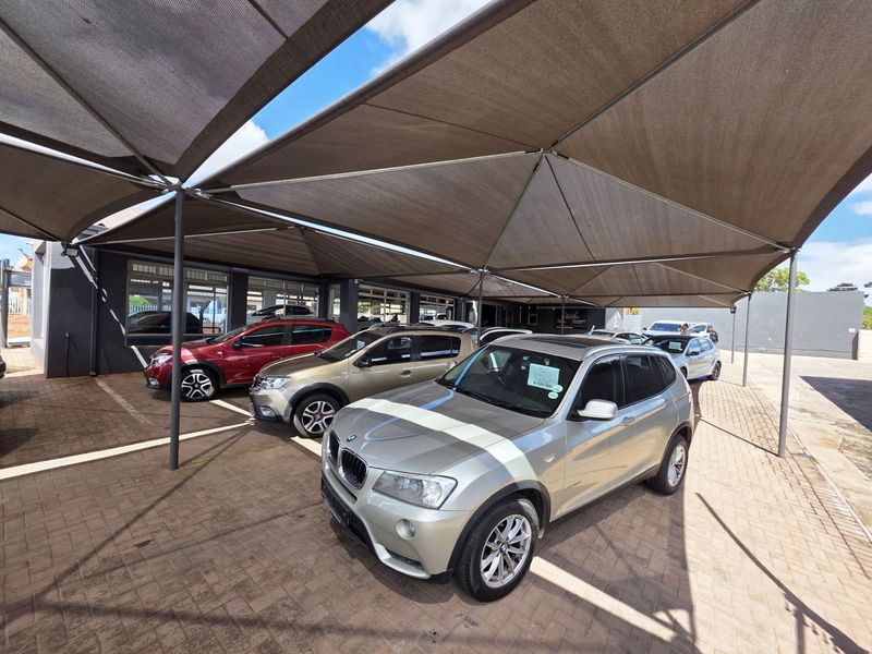 A paved outdoor vehicle display area featuring a large, structured shade cloth canopy supported by steel columns.