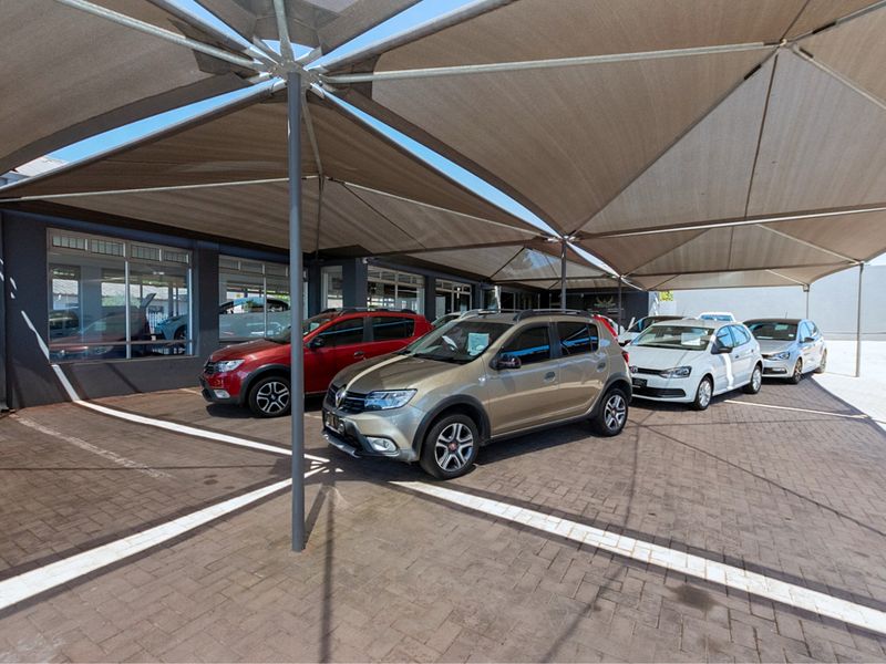 A well-maintained retail display area featuring a large shade sail structure over a paved parking lot with expansive glass frontage.