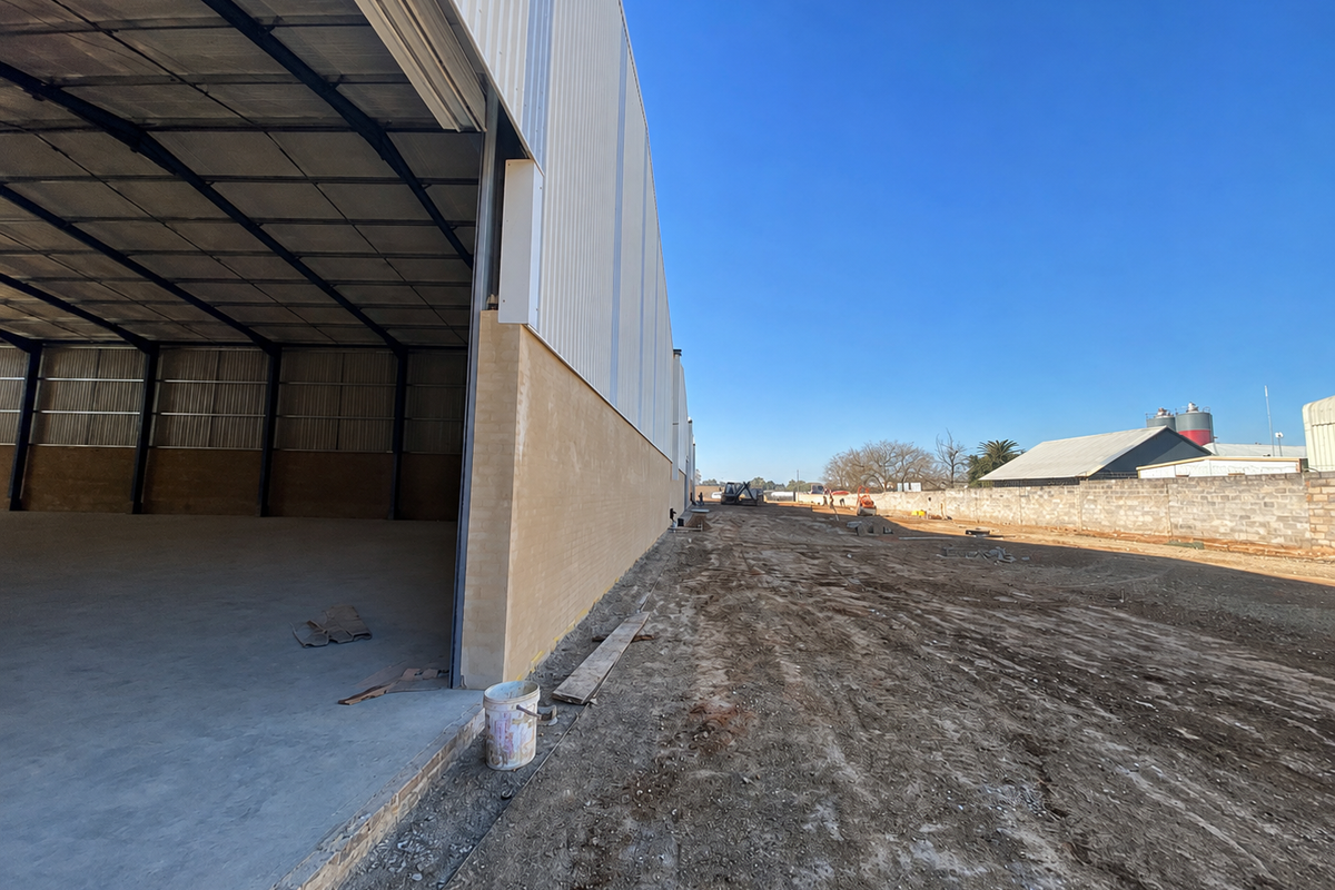 Exterior view of an industrial warehouse facility featuring a large open bay, steel portal frame construction, and an expansive unpaved yard area.