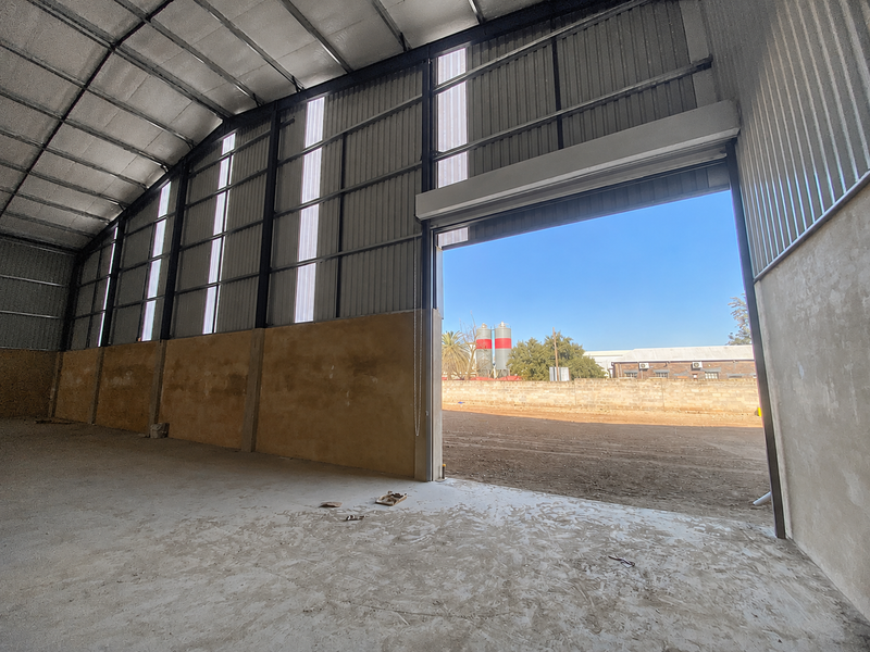 Spacious industrial warehouse unit featuring a high-clearance corrugated metal roof, concrete flooring, and a large roller shutter door for vehicle access.