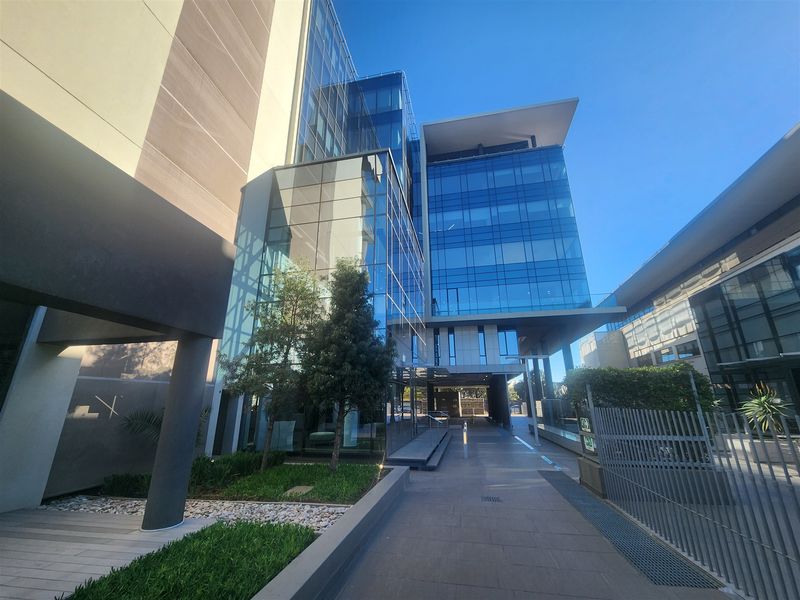 Modern commercial office building featuring a glass curtain wall facade, paved pedestrian walkway, and landscaped garden areas.