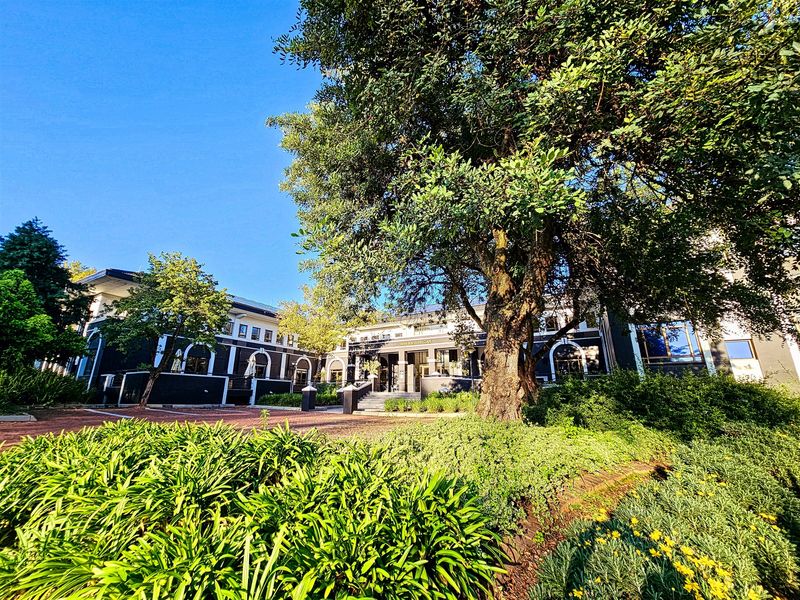 A well-maintained office building exterior featuring a landscaped garden, mature trees, and a paved driveway leading to the main entrance.