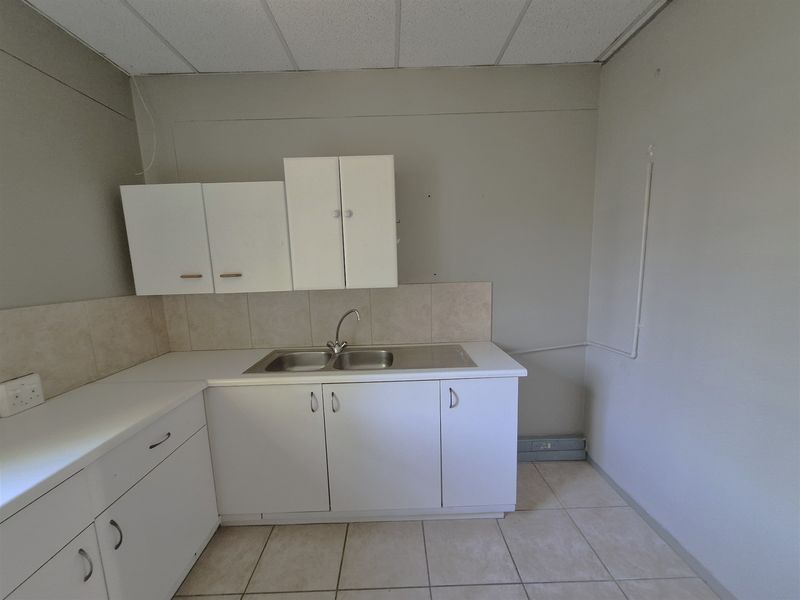 A functional office kitchenette featuring white built-in cabinetry, a stainless steel sink, tiled backsplash, and durable tiled flooring.