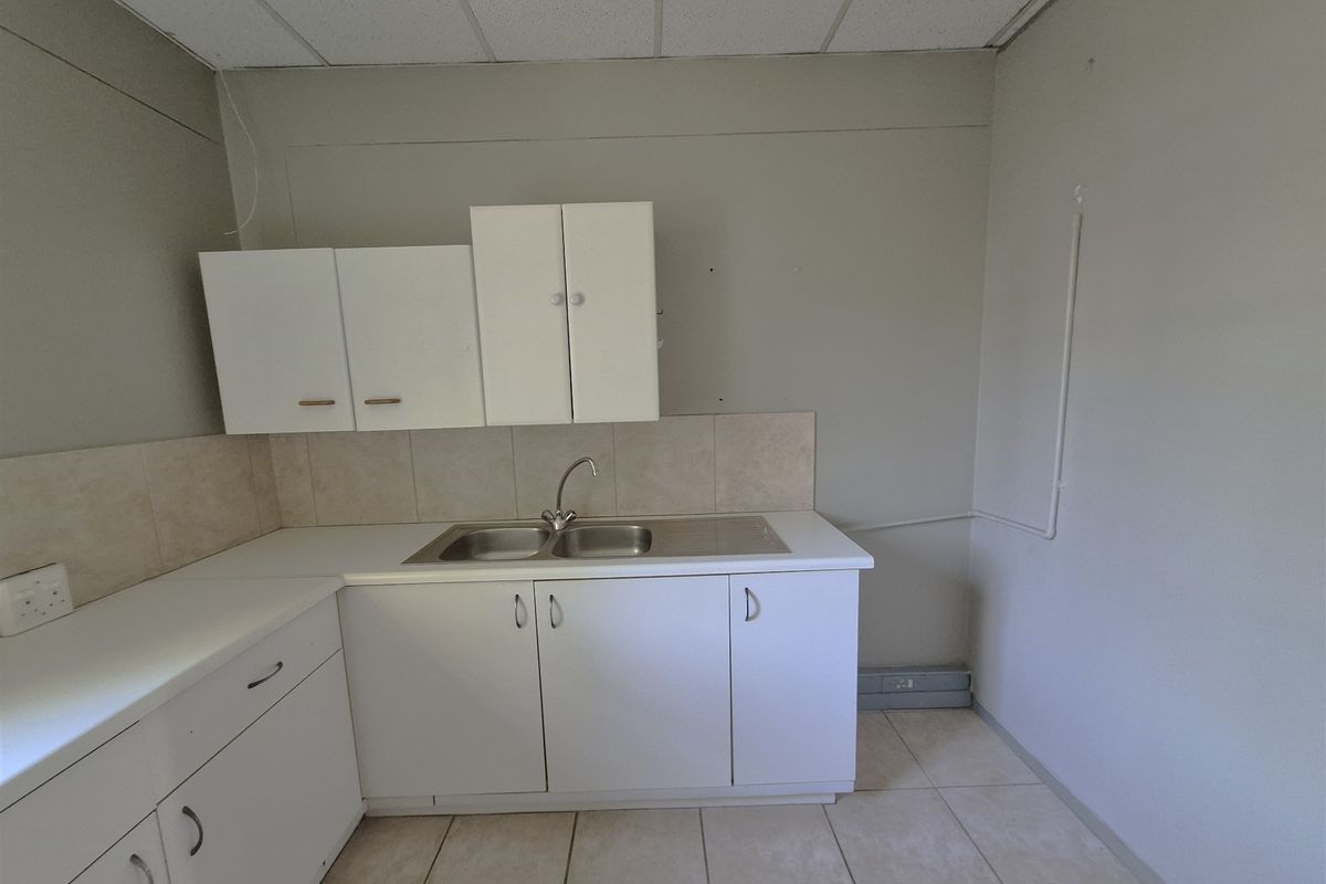 A functional office kitchenette featuring white built-in cabinetry, a stainless steel sink, tiled backsplash, and durable tiled flooring.