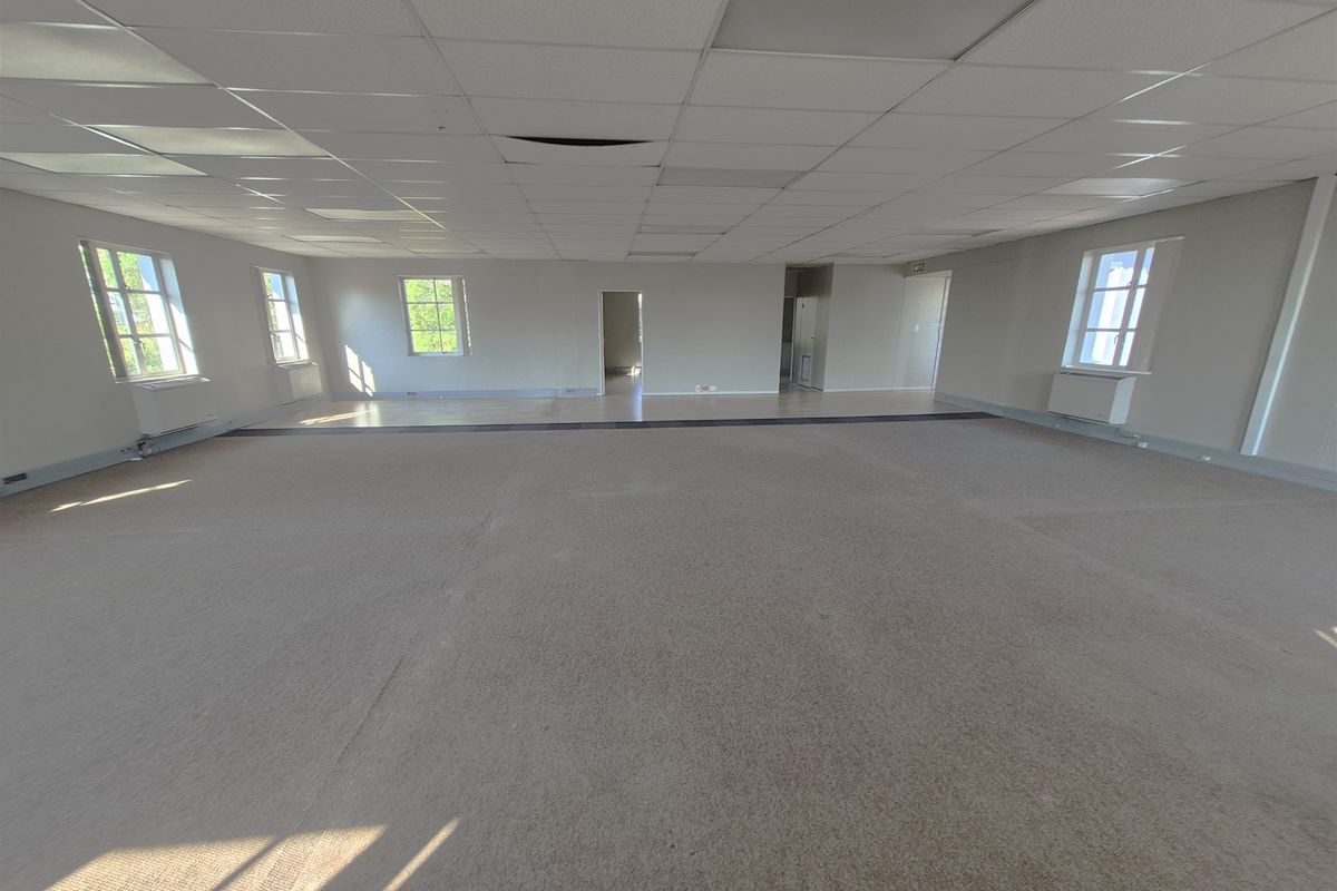 A spacious, vacant open-plan office area featuring a suspended ceiling, carpeted flooring, and multiple windows providing natural light.