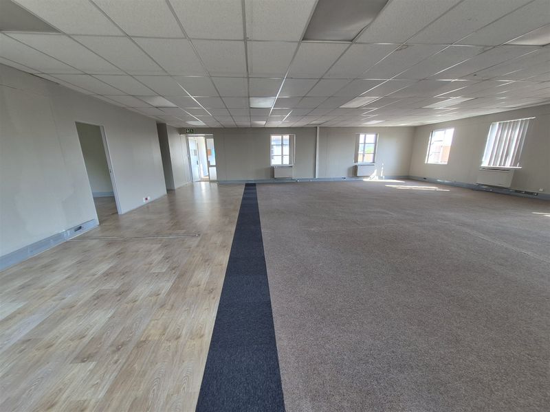 A spacious, empty open-plan office area featuring a mix of laminate and carpet flooring, suspended ceilings, and wall-mounted HVAC units.