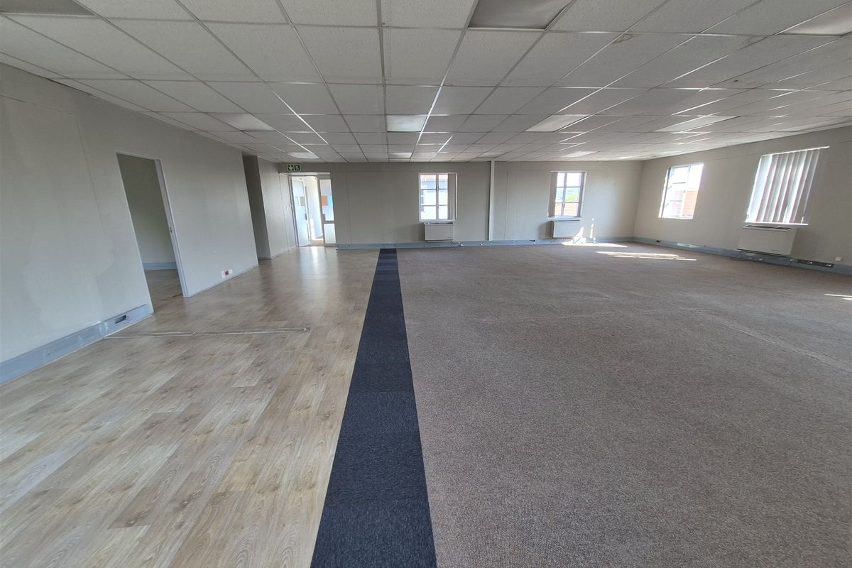 A spacious, empty open-plan office area featuring a mix of laminate and carpet flooring, suspended ceilings, and wall-mounted HVAC units.