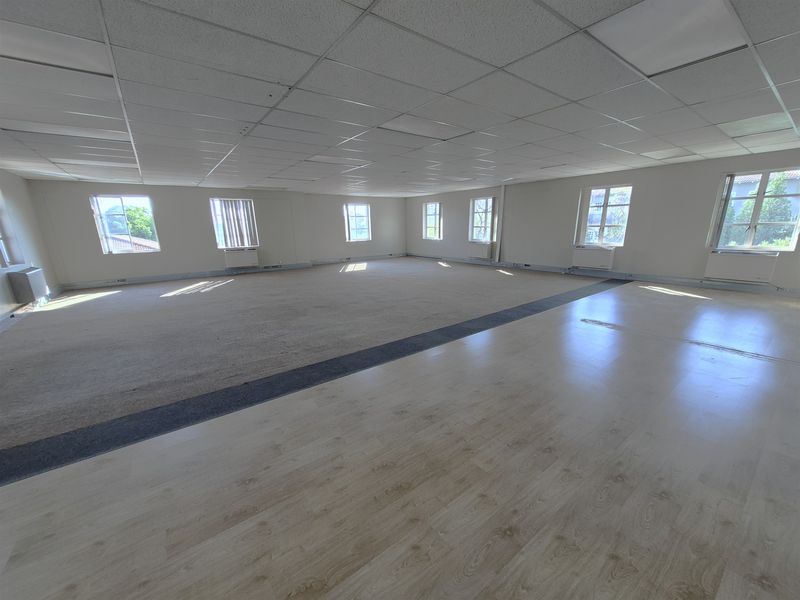 Spacious open-plan office area featuring a mix of laminate and carpet flooring, suspended ceiling tiles, and multiple windows providing natural light.