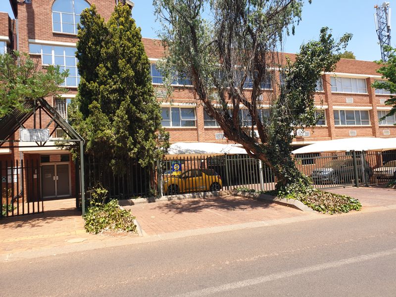 Front exterior view of a multi-story brick office building featuring paved parking, shade cloth carports, and secure perimeter fencing.