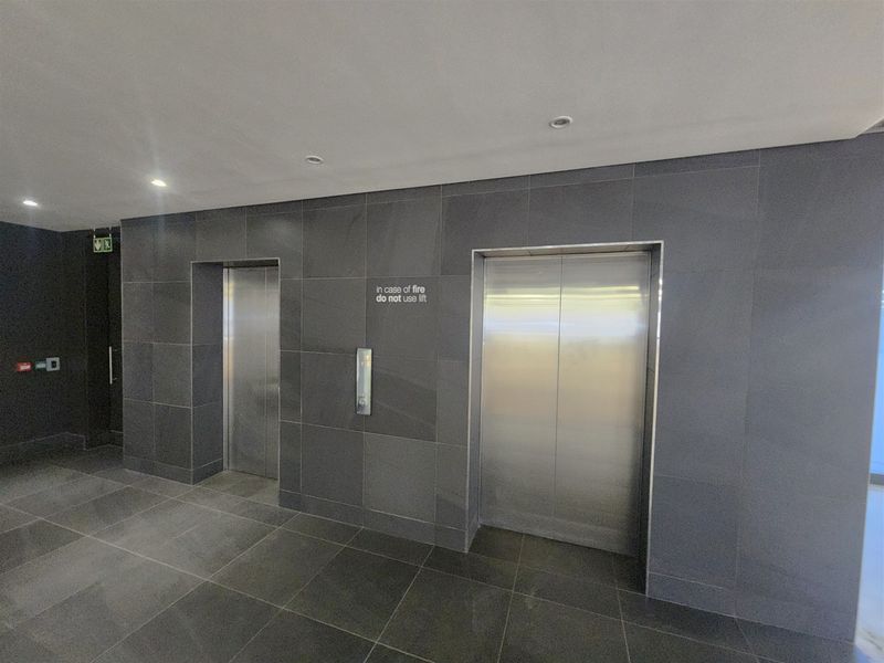 Modern office building elevator lobby featuring sleek stainless steel lift doors and contemporary dark grey tiled wall and floor finishes.