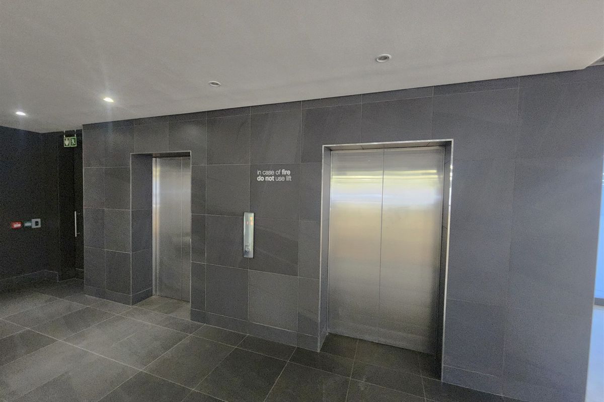 Modern office building elevator lobby featuring sleek stainless steel lift doors and contemporary dark grey tiled wall and floor finishes.
