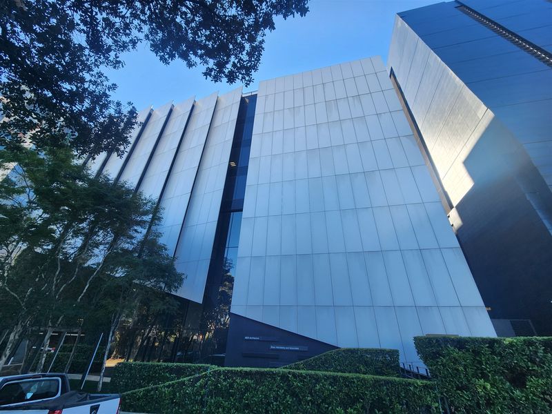 A modern office building facade featuring vertical cladding panels, extensive glass curtain walling, and manicured perimeter hedging.