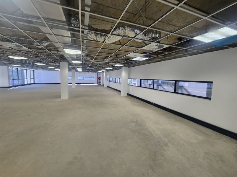 A large, open-plan office space featuring an unfinished ceiling with exposed ducting, structural columns, and horizontal strip windows, ready for custom fit-out.