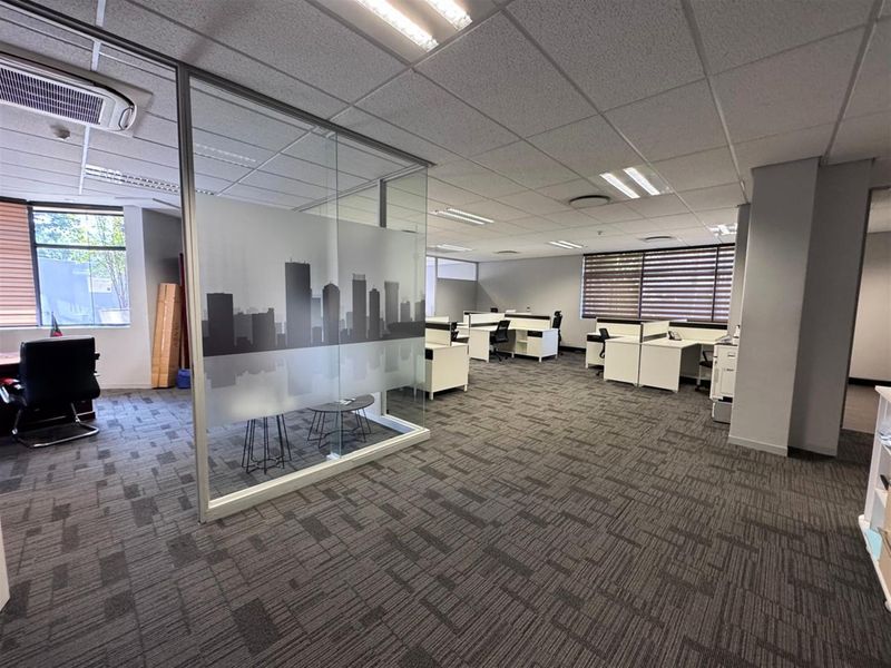 Open-plan office space featuring glass partitioning, carpeted flooring, suspended ceilings with recessed lighting, and large windows with venetian blinds.