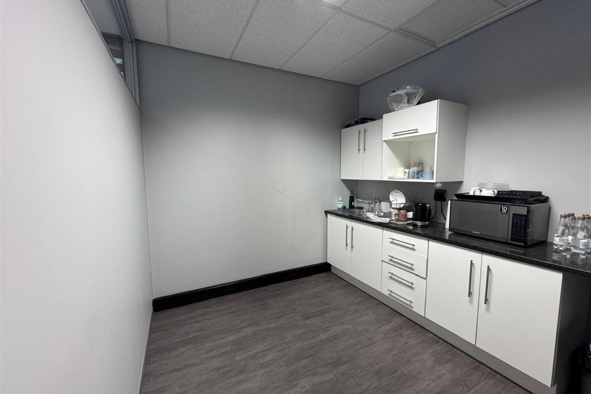A functional office kitchenette featuring white built-in cabinetry, dark laminate countertops, and durable vinyl flooring under a drop ceiling.