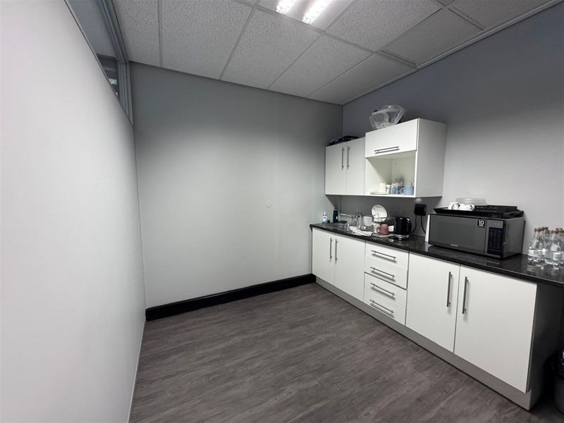 A functional office kitchenette featuring white built-in cabinetry, dark laminate countertops, and durable vinyl flooring under a drop ceiling.