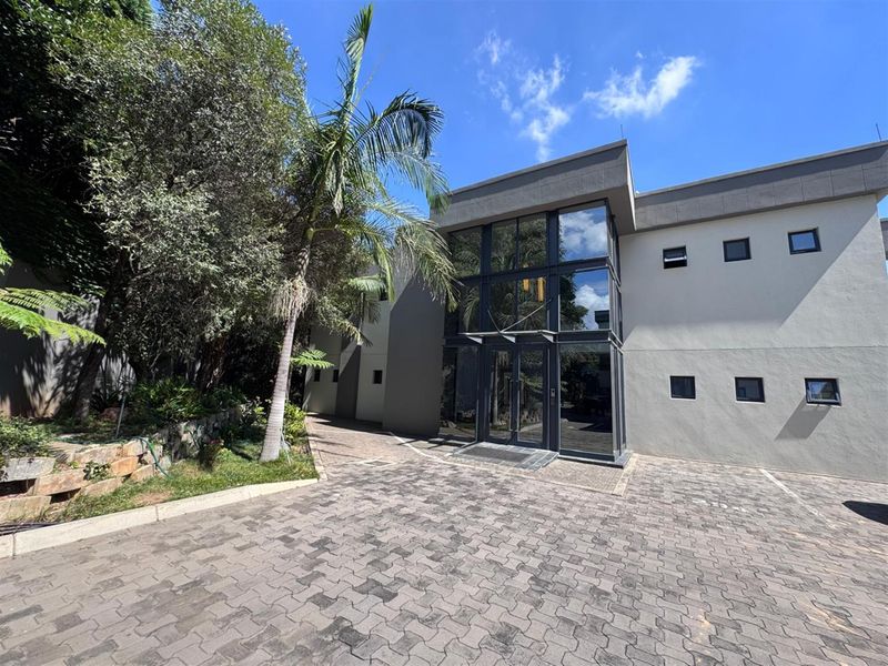 Modern office building exterior featuring a prominent glass facade, double-height entrance, and a paved driveway with surrounding landscaped garden.