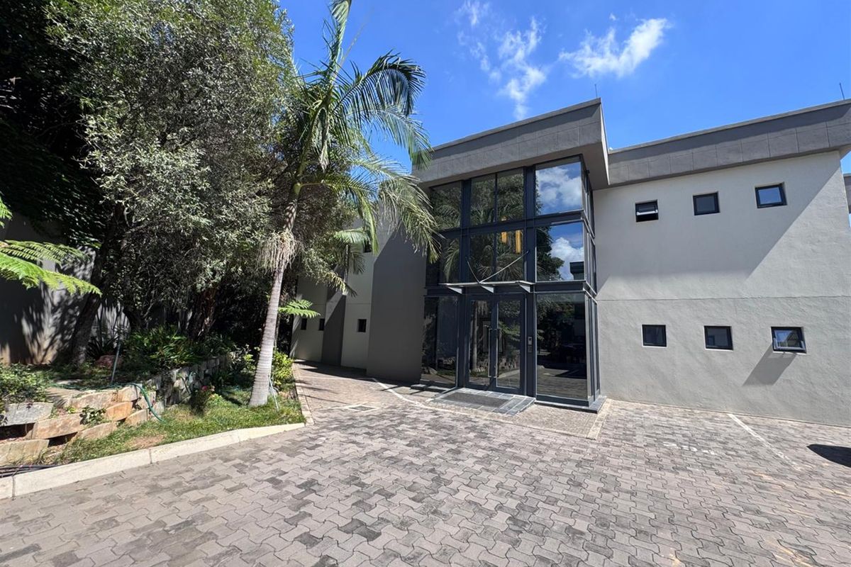 Modern office building exterior featuring a prominent glass facade, double-height entrance, and a paved driveway with surrounding landscaped garden.