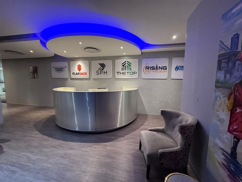 Modern office reception area featuring a curved metallic desk, recessed ceiling lighting with blue accent cove lighting, and wood-look laminate flooring.