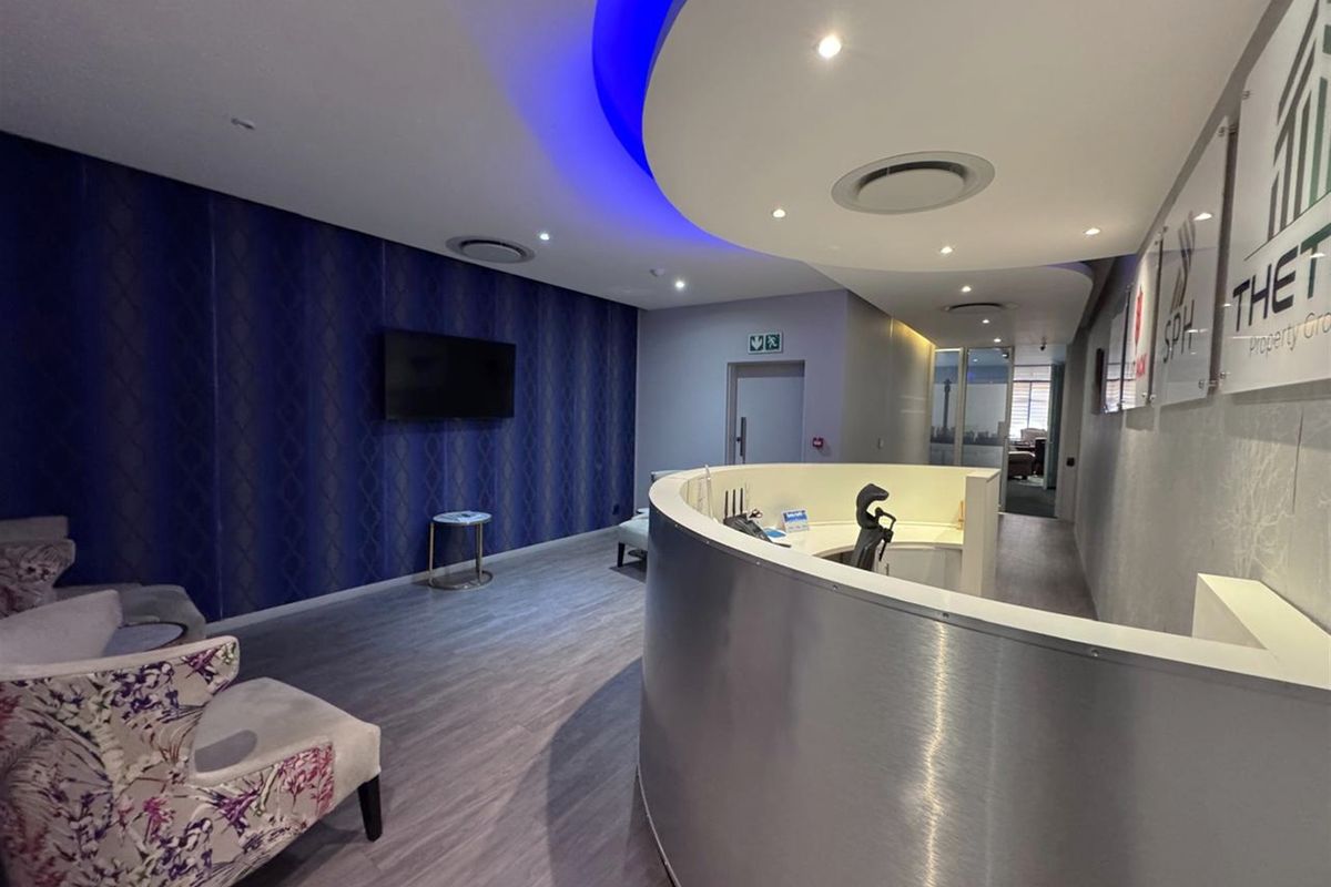 Modern office reception area featuring a curved metallic reception desk, decorative wallpaper, and integrated recessed ceiling lighting with blue accent cove lighting.