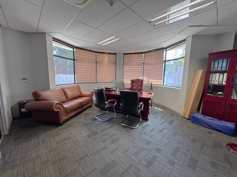 A professional office space featuring a curved window wall with fitted blinds, suspended ceiling tiles, and carpeted flooring.
