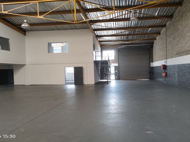 Spacious industrial warehouse unit featuring high ceilings with exposed steel trusses, concrete flooring, a large roller shutter door, and an internal staircase.