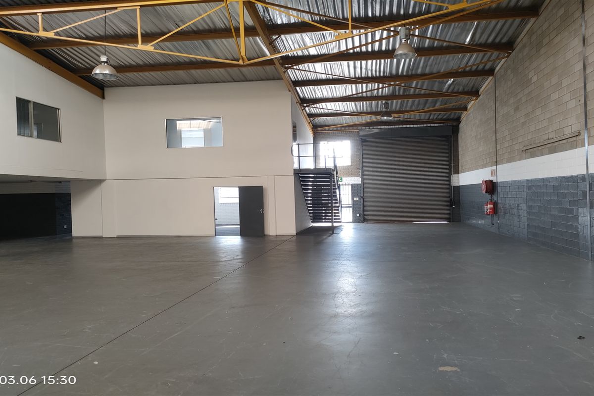 Spacious industrial warehouse unit featuring high ceilings with exposed steel trusses, concrete flooring, a large roller shutter door, and an internal staircase.