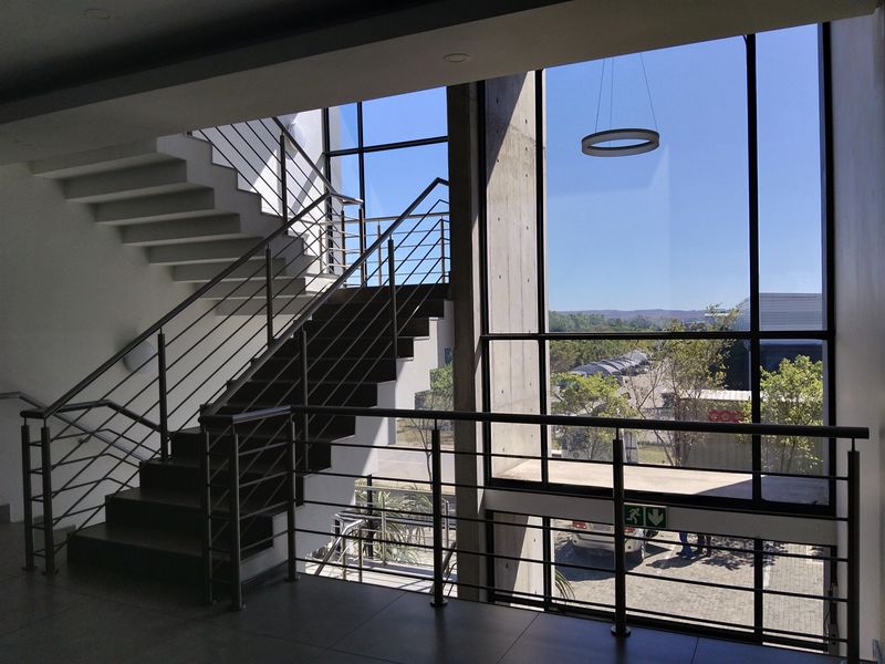 Modern industrial hallway featuring a concrete staircase with stainless steel railings and expansive floor-to-ceiling windows offering natural light and exterior views.
