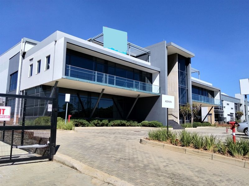 Modern industrial office building featuring a glass facade, balconies, and a paved entrance area with secure gated access.
