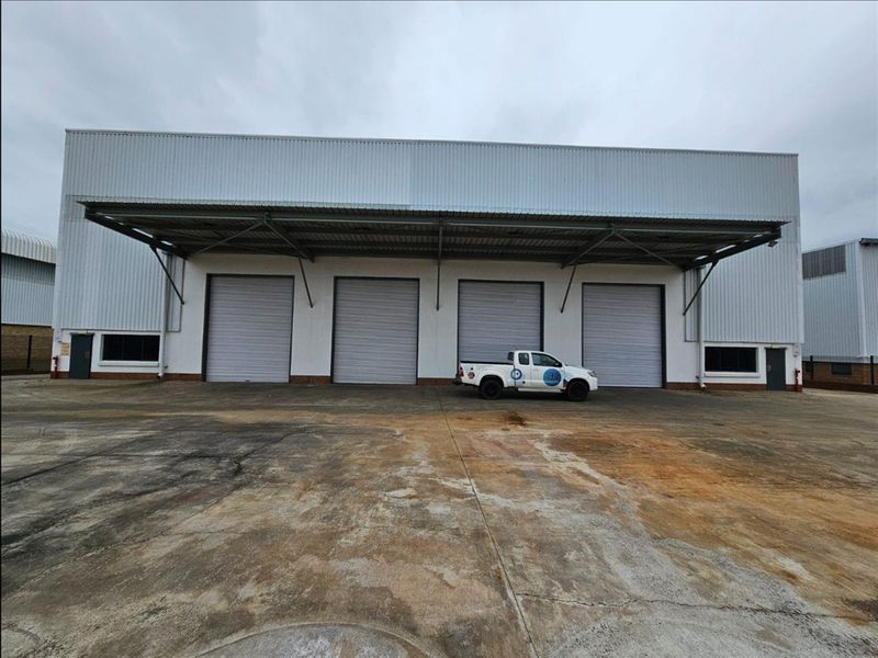 Industrial warehouse exterior featuring four large roller shutter doors, a covered loading canopy, and a spacious concrete hardstand area.