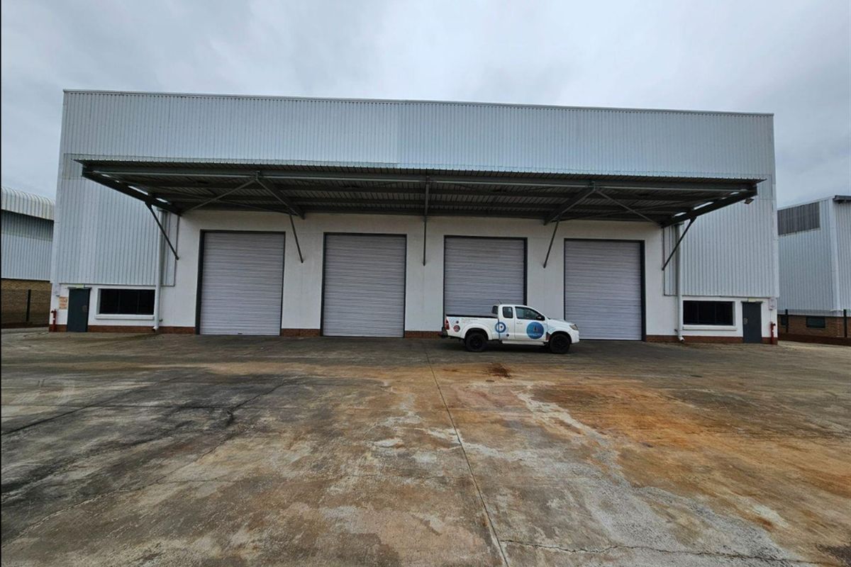 Industrial warehouse exterior featuring four large roller shutter doors, a covered loading canopy, and a spacious concrete hardstand area.