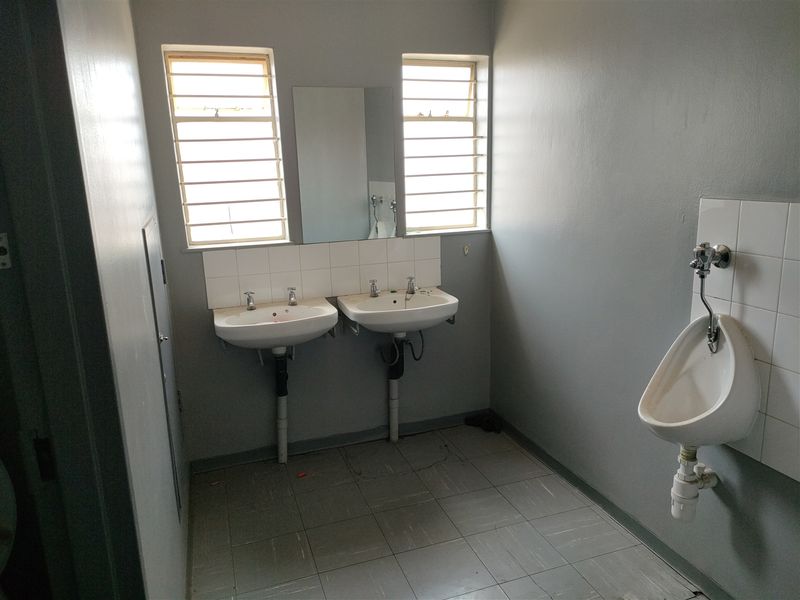A functional industrial-style bathroom featuring dual washbasins, a wall-mounted urinal, and louvered windows, currently in a basic, dated condition.