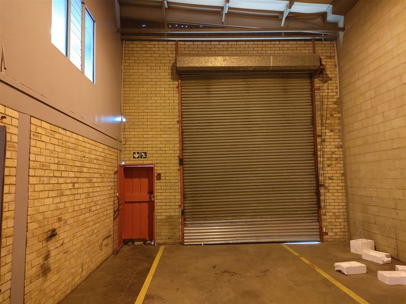 Industrial warehouse space featuring a large roller shutter door, exposed brick walls, concrete flooring, and an emergency exit door.