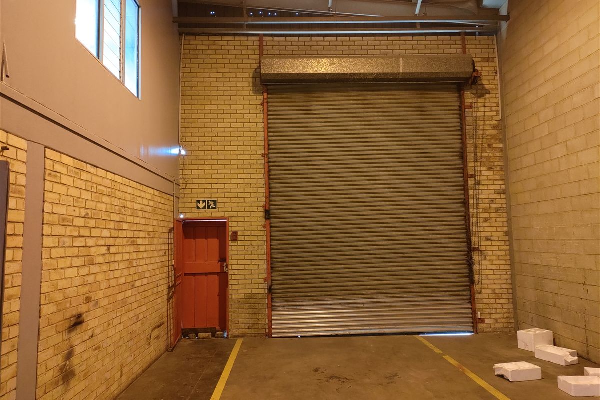 Industrial warehouse space featuring a large roller shutter door, exposed brick walls, concrete flooring, and an emergency exit door.