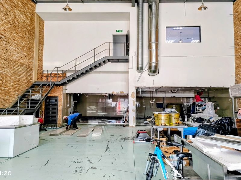 Spacious industrial unit featuring exposed brick walls, a metal staircase to a mezzanine level, and high-capacity ventilation ducting, currently undergoing renovation.