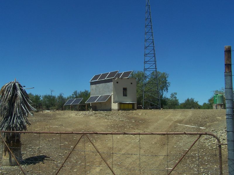 A rural farm utility building featuring a solar panel array and a tall communication tower, set on a dry, open landscape.
