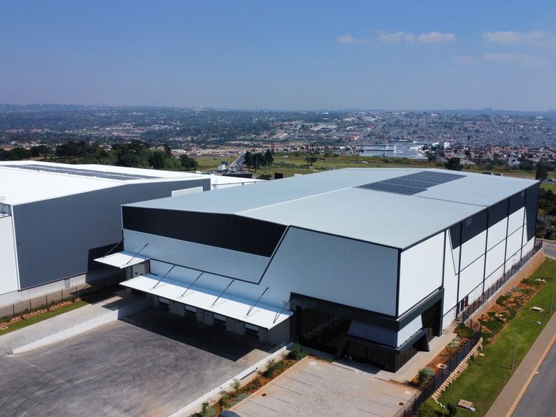 Aerial view of a modern industrial warehouse facility featuring a large metal roof with solar panels, loading docks, and an expansive asphalt yard.