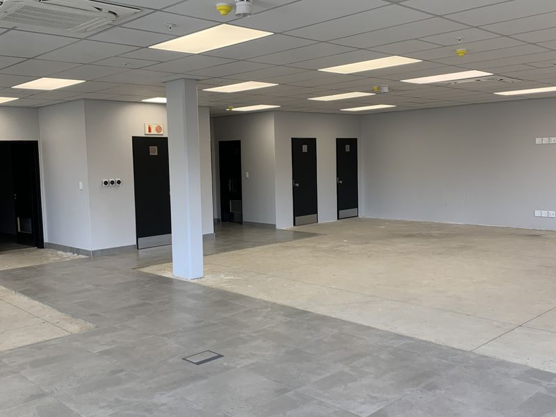 A spacious, open-plan industrial office area featuring a suspended ceiling, recessed lighting, air conditioning, and a mix of tiled and concrete flooring.